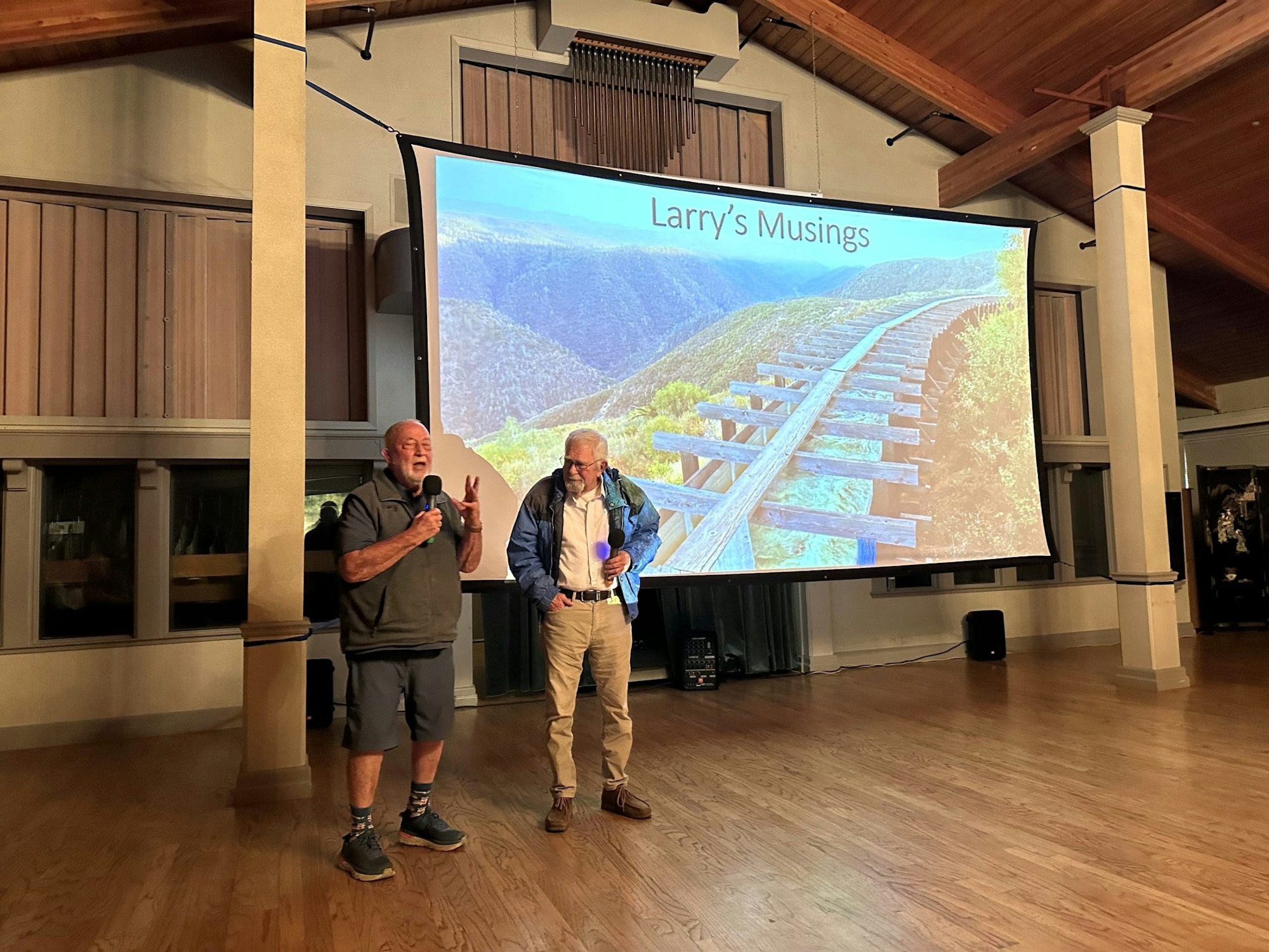 Two people are speaking on stage with a projection titled "Larry's Musings" showing a mountain landscape.