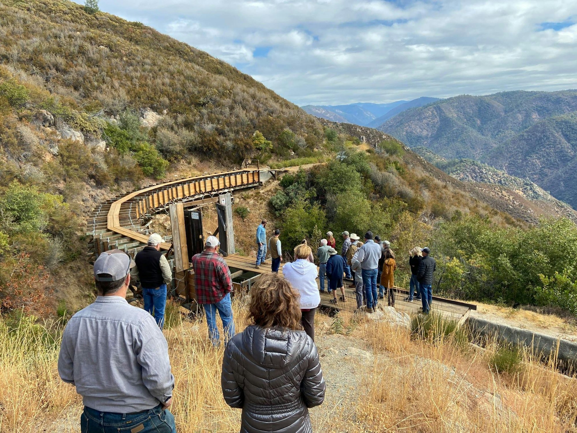 A group of people touring a wooden walkway on a hillside, with scenic mountain views in the background.
