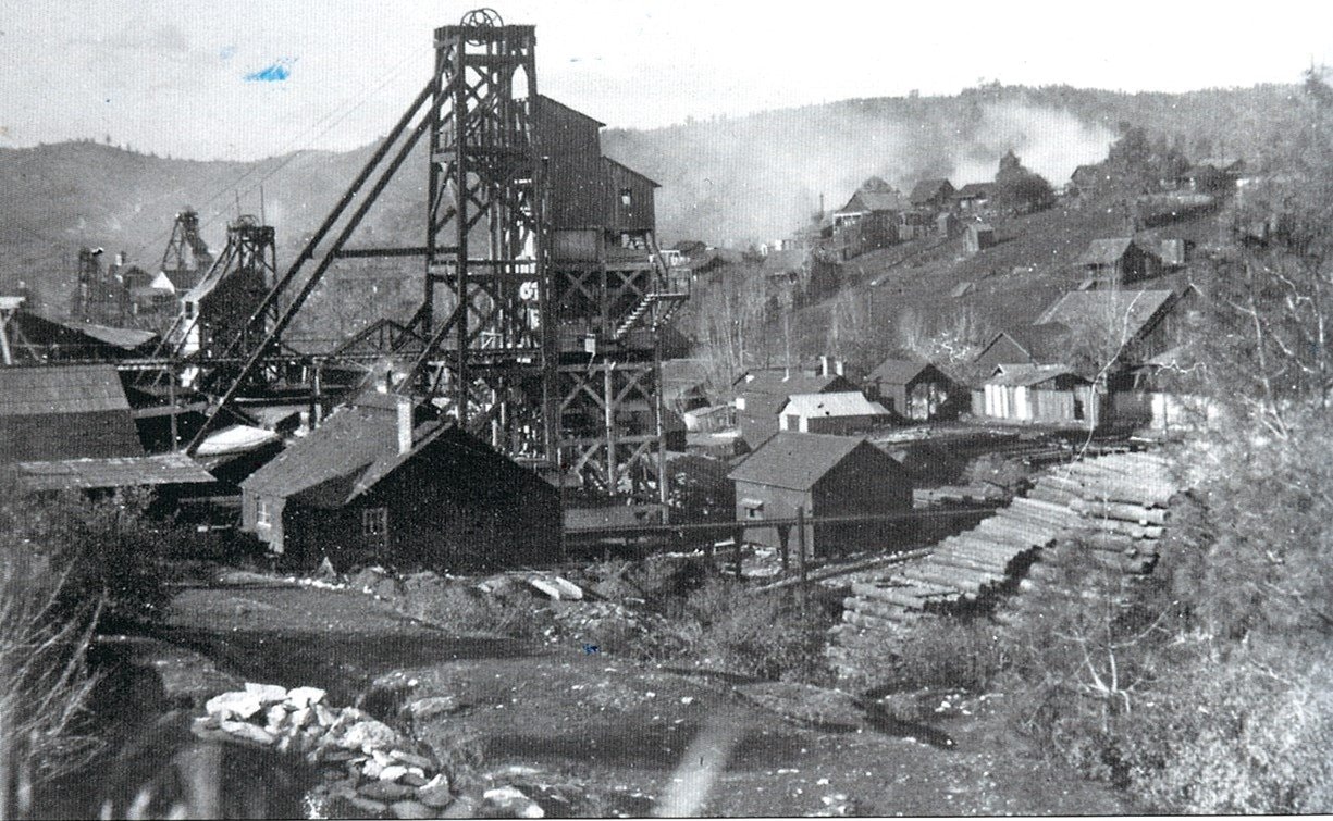 A black-and-white image of an old mining site with structures, houses, and stacked logs in a hilly landscape.