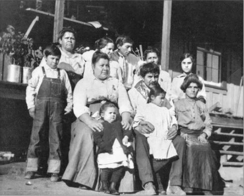 A group portrait of eleven people, including men, women, and children, posed in front of a wooden building.