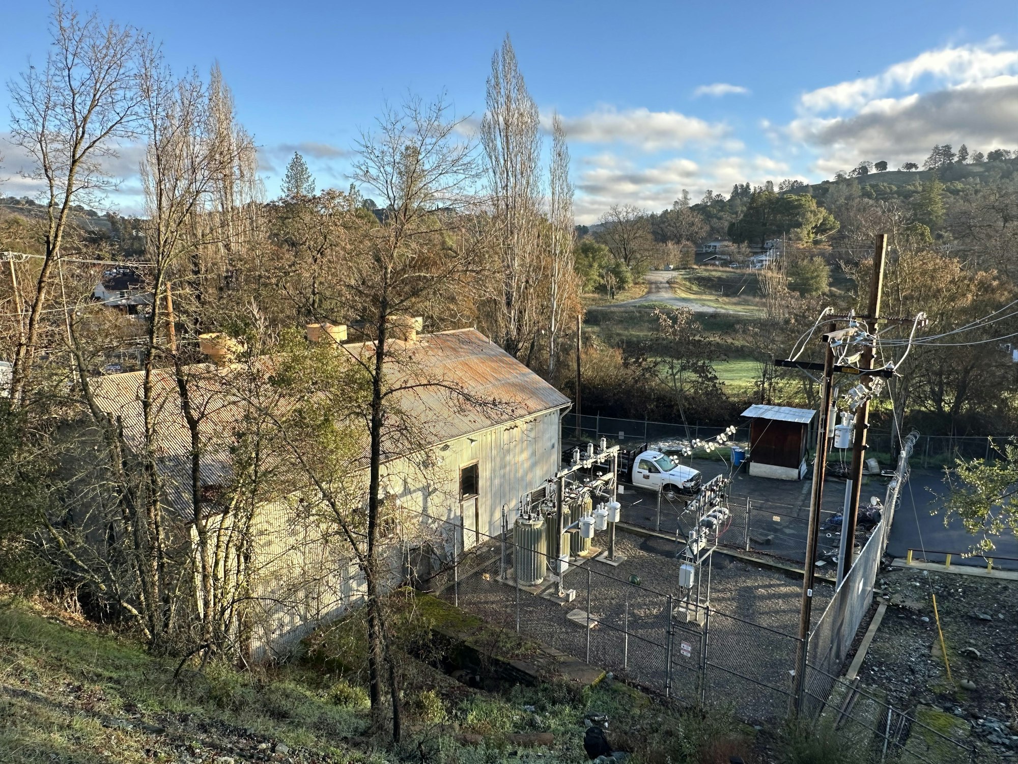 A small electrical substation next to a weathered building, surrounded by trees and hills under a clear sky.