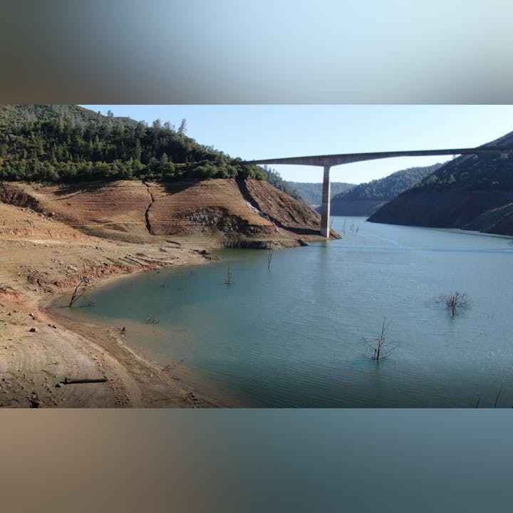 A reservoir with low water levels, exposed land, and a bridge crossing over it, surrounded by hills and sparse trees.