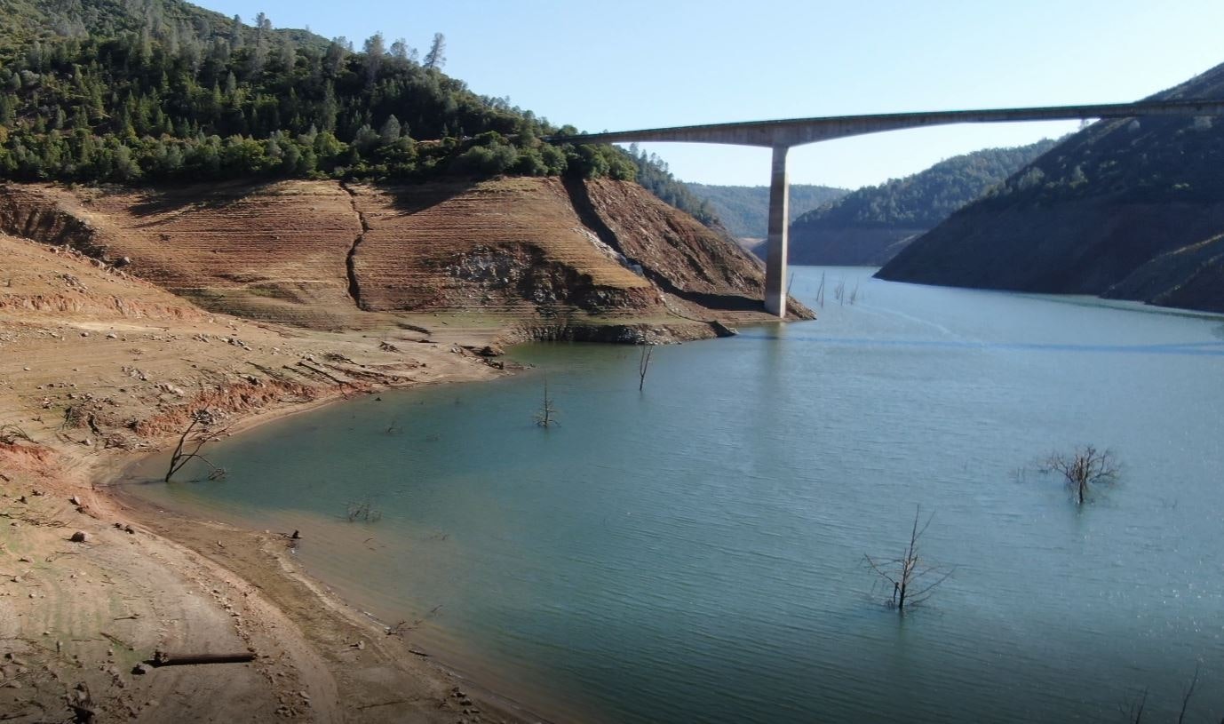 A reservoir with low water levels, exposed land, and a bridge crossing over it, surrounded by hills and sparse trees.