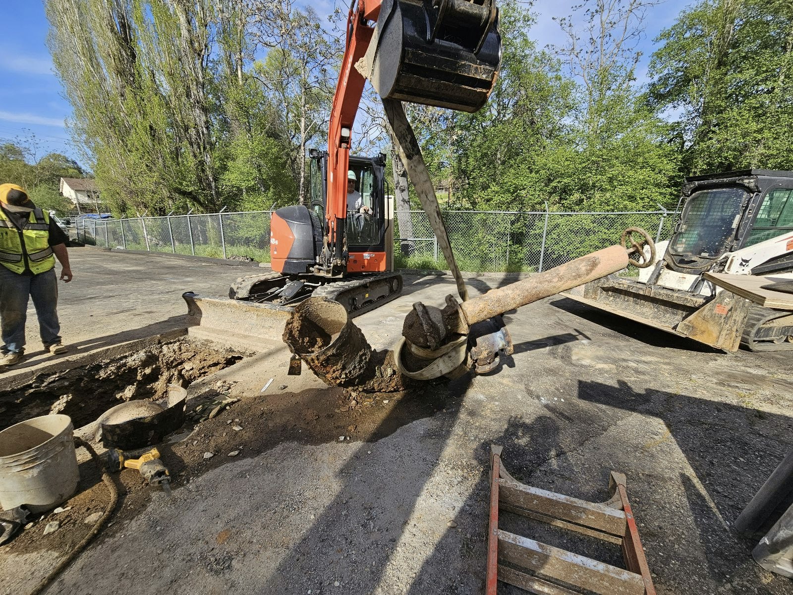 Construction site with an excavator lifting a large rusty valve, a worker nearby, and various equipment around.