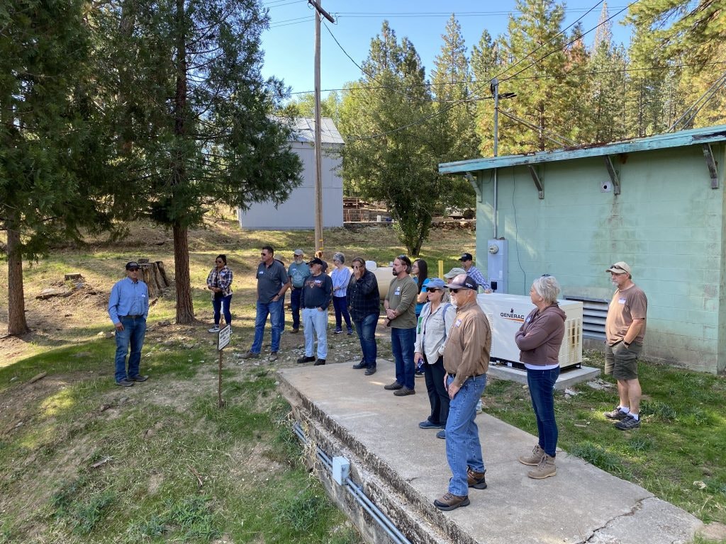 A group of people standing outdoors near a generator and building, listening to a person on the left. Trees and grass in the background.