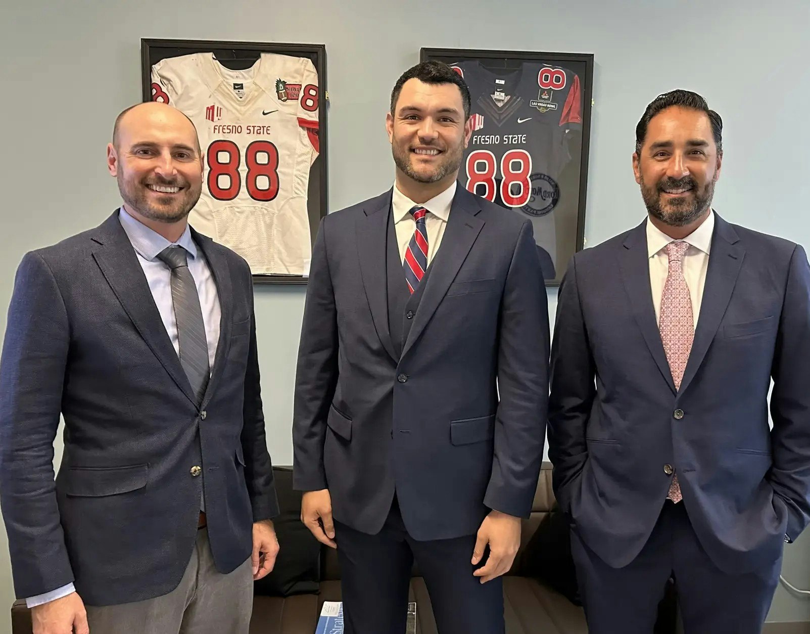 Three men in suits standing in front of framed Fresno State jerseys.