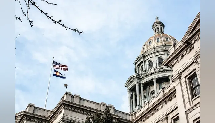 A domed government building with the US and Colorado flags flying against a cloudy sky.