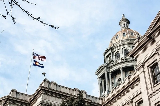 A domed government building with the US and Colorado flags flying against a cloudy sky.