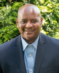A smiling man in a suit with a light blue shirt, standing outdoors with greenery in the background.