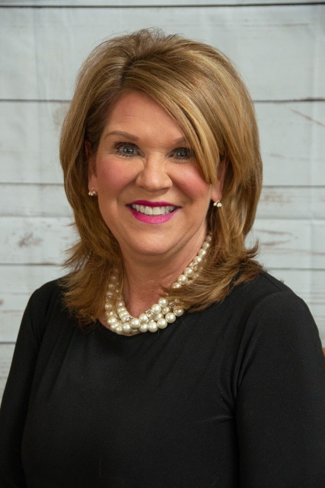 A person with blonde hair, wearing a black top and pearl necklace, smiles against a wooden panel background.