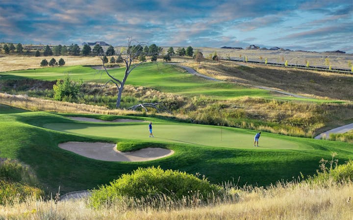 A scenic golf course with two players, rolling hills, green fairways, sand bunkers, and a distant building under a cloudy sky.