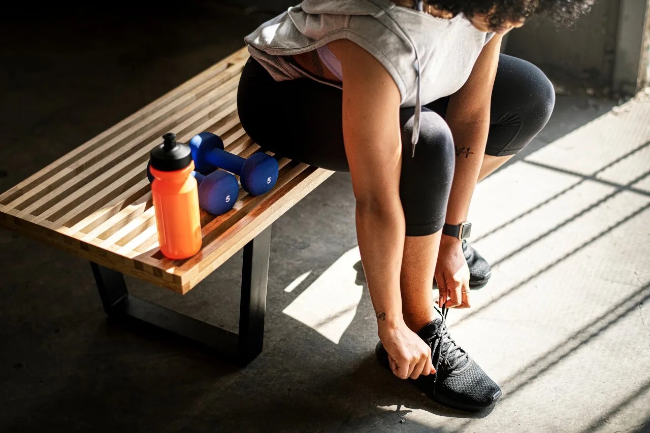 Person tying shoe near water bottle and dumbbells on bench, suggesting a workout setting.