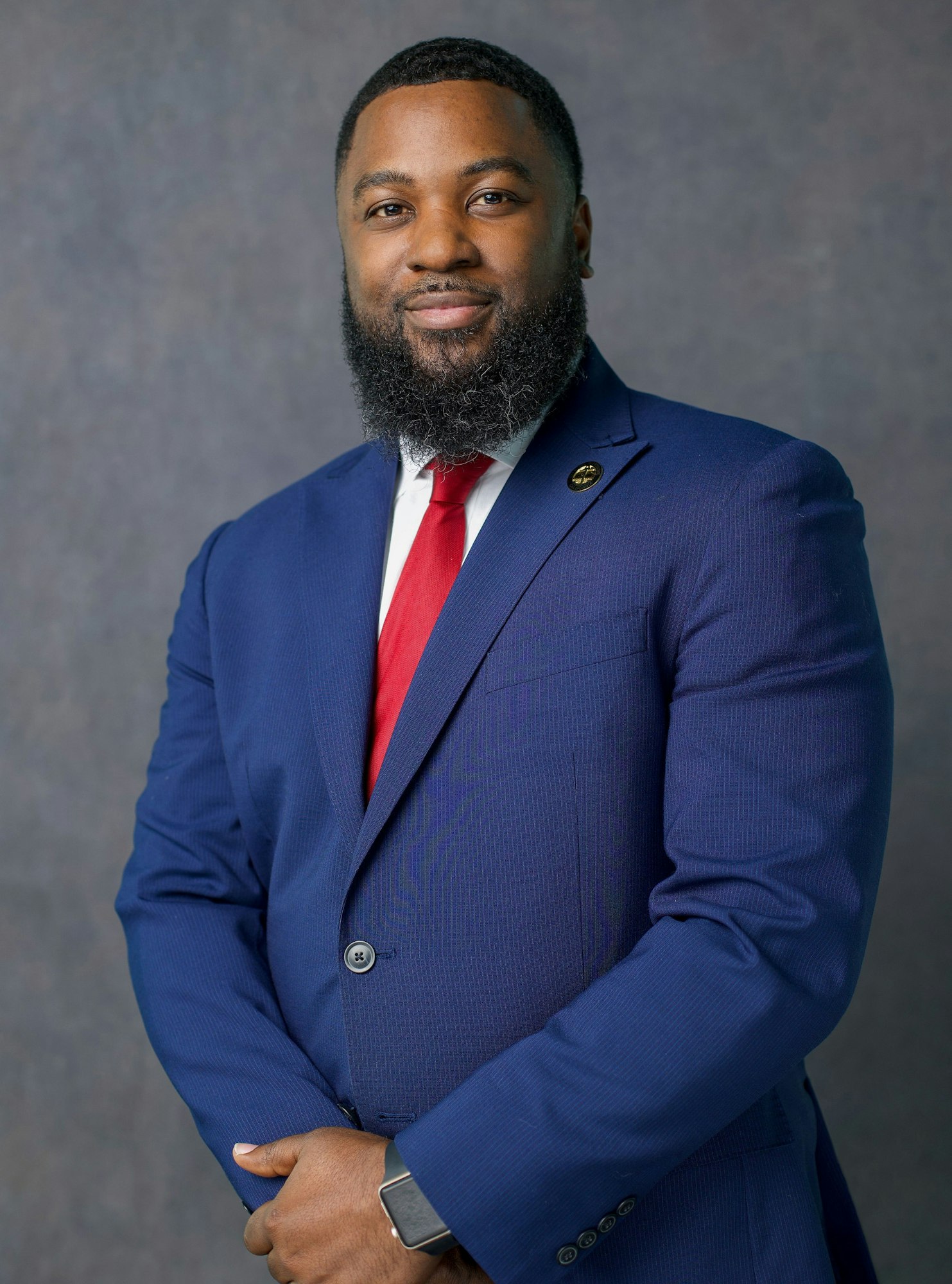 A man wearing a blue suit and red tie, posing against a gray background.