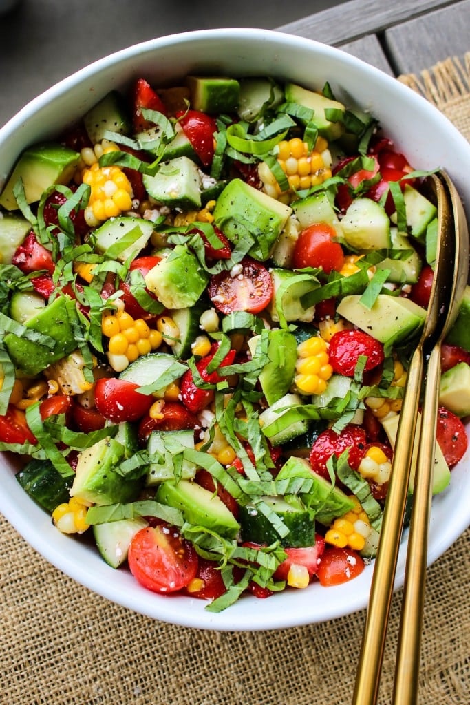 A colorful salad with avocado, cherry tomatoes, cucumber, corn, and greens in a white bowl with golden utensils.