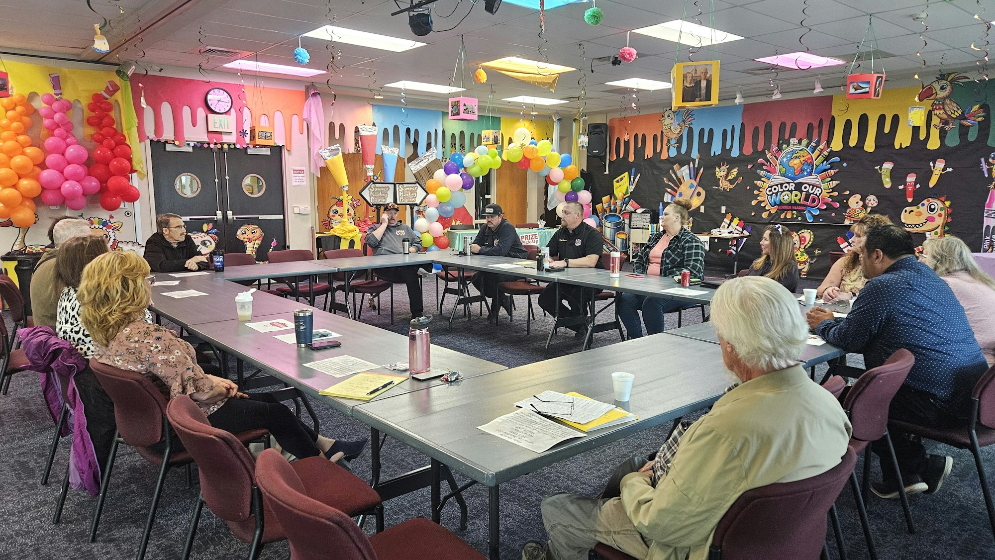 A group of people in a colorfully decorated room sit around a U-shaped table, discussing or holding a meeting.