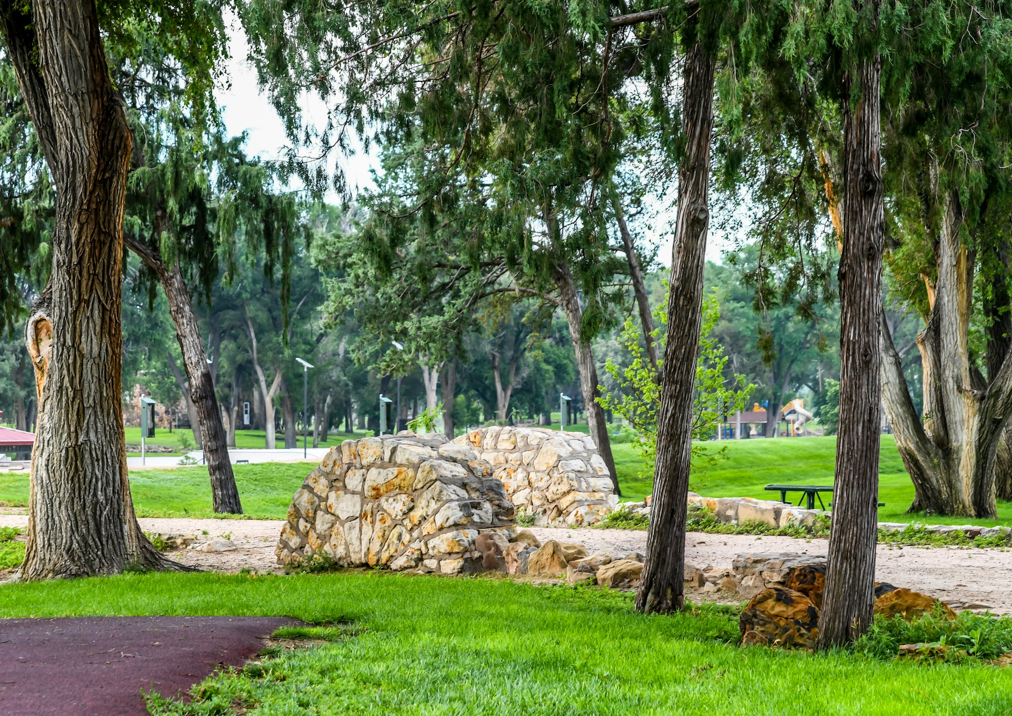 A peaceful park scene with trees, grass, a pathway, stone formations, and a picnic table.