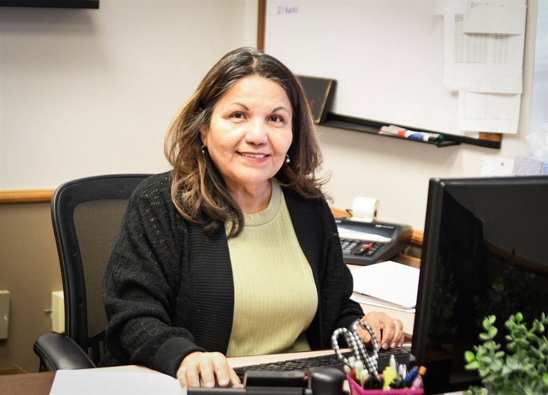 A smiling woman at a desk with a computer, phone, and office supplies.