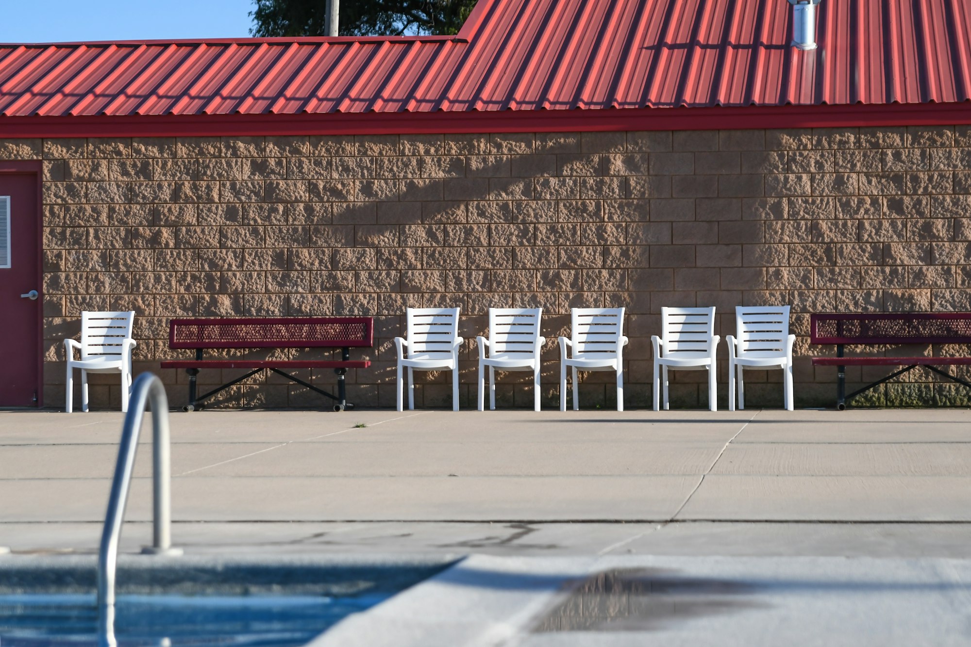 A poolside area with white chairs, benches, and a brick building with a red roof in the background.