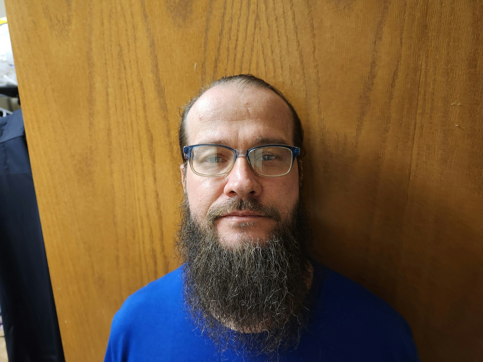 A bearded man with glasses standing against a wooden background, wearing a blue shirt.