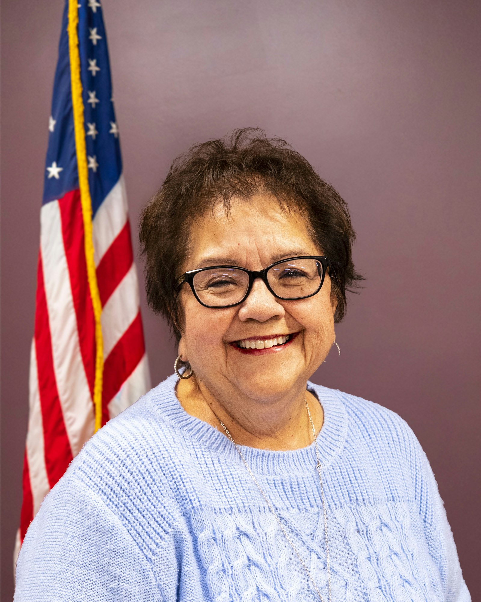 A smiling individual in a light blue sweater stands in front of an American flag against a purple background.