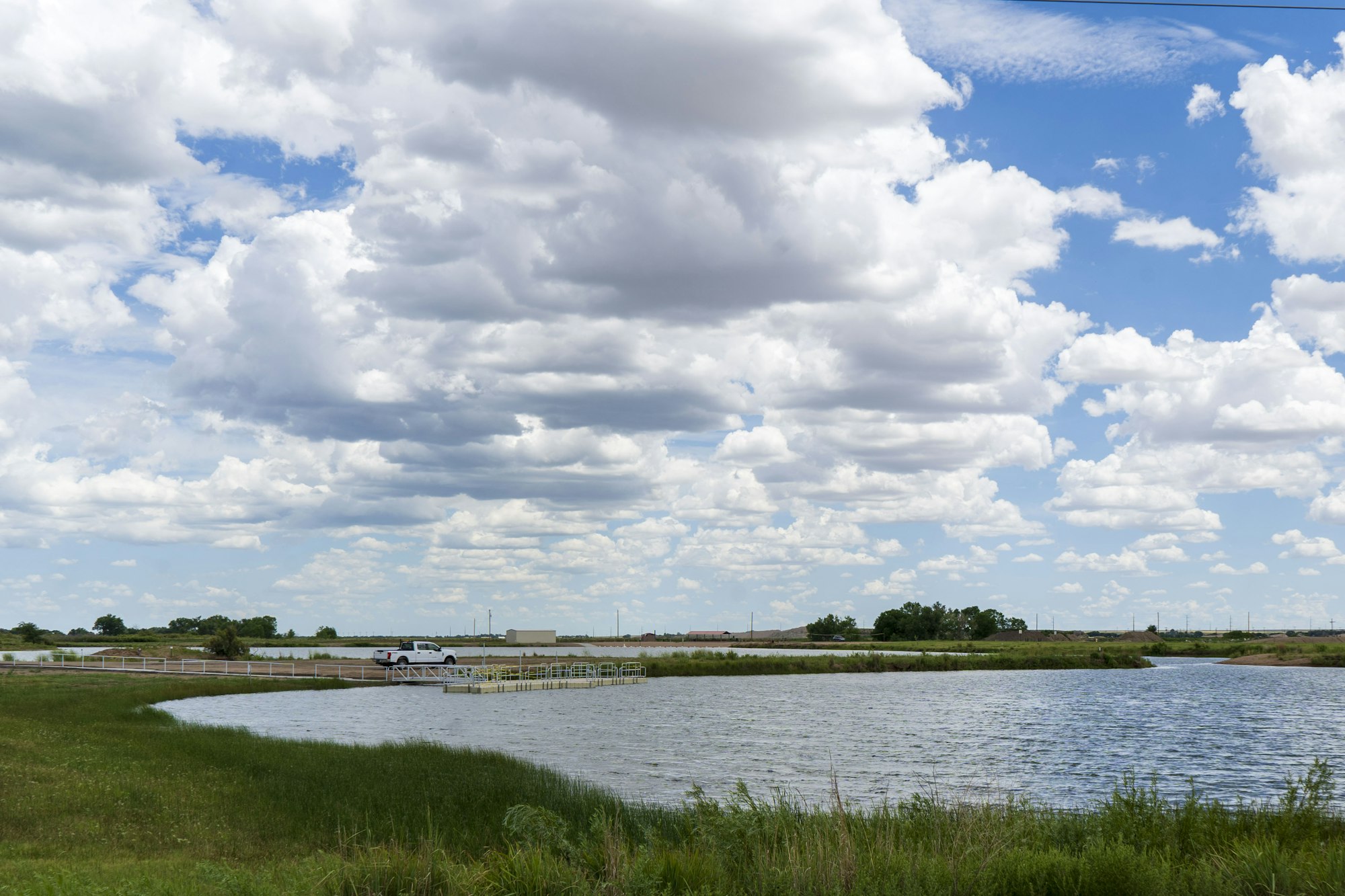 A truck by a lake under a big, partly cloudy sky with grass and a pier.