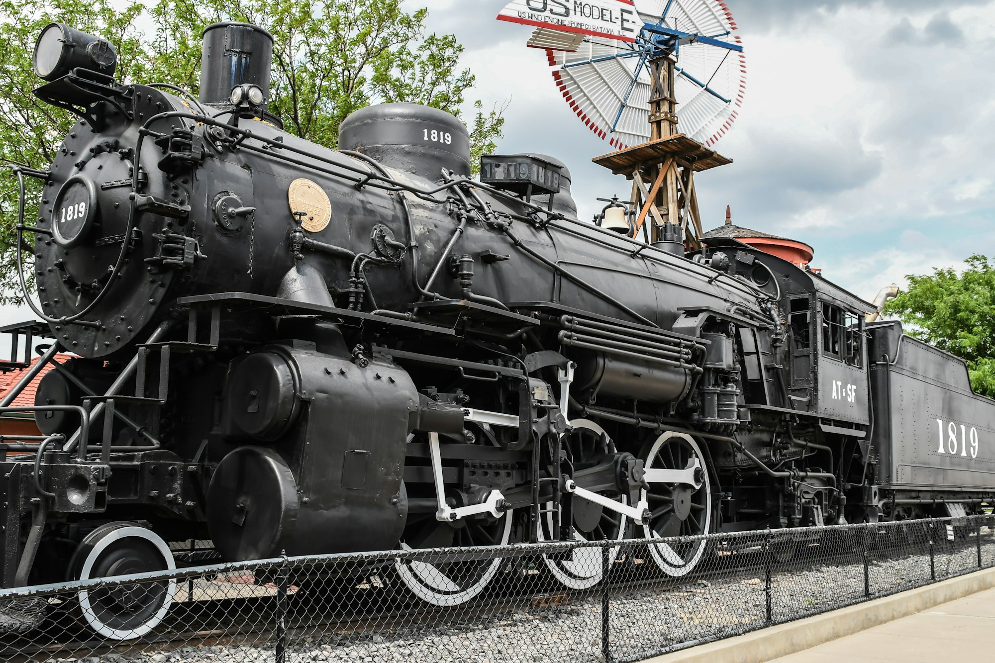 Old steam locomotive 1819 displayed outdoors near a windmill, behind a chain-link fence.