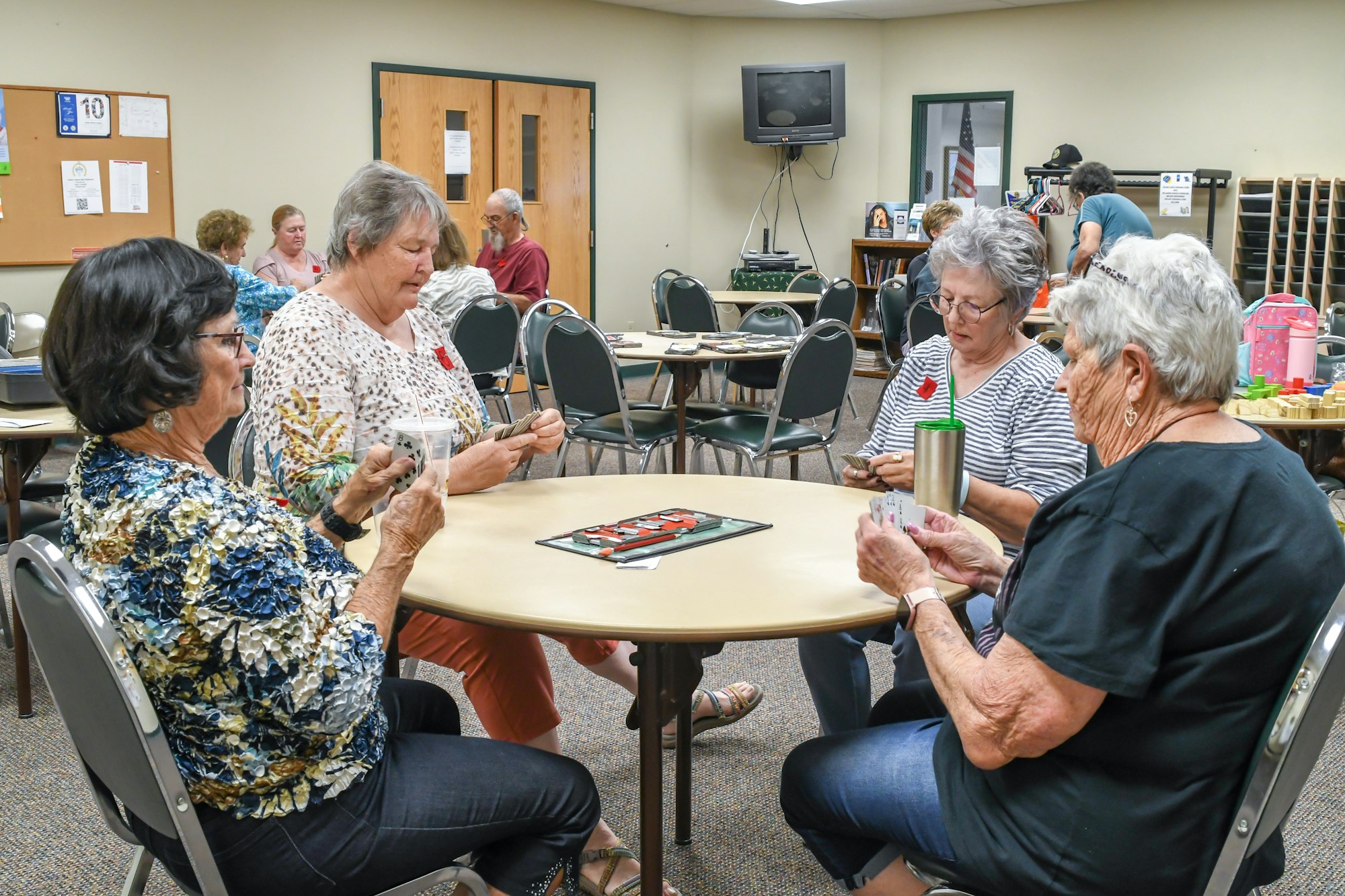 Four people playing cards at a round table in a room with others.