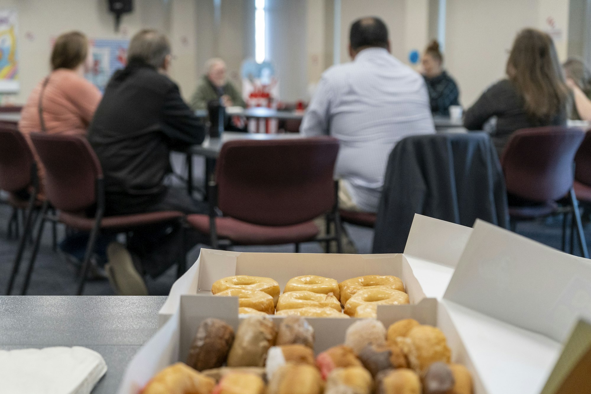 A box of donuts is on a table in the foreground, with a group of people sitting and chatting in the background.