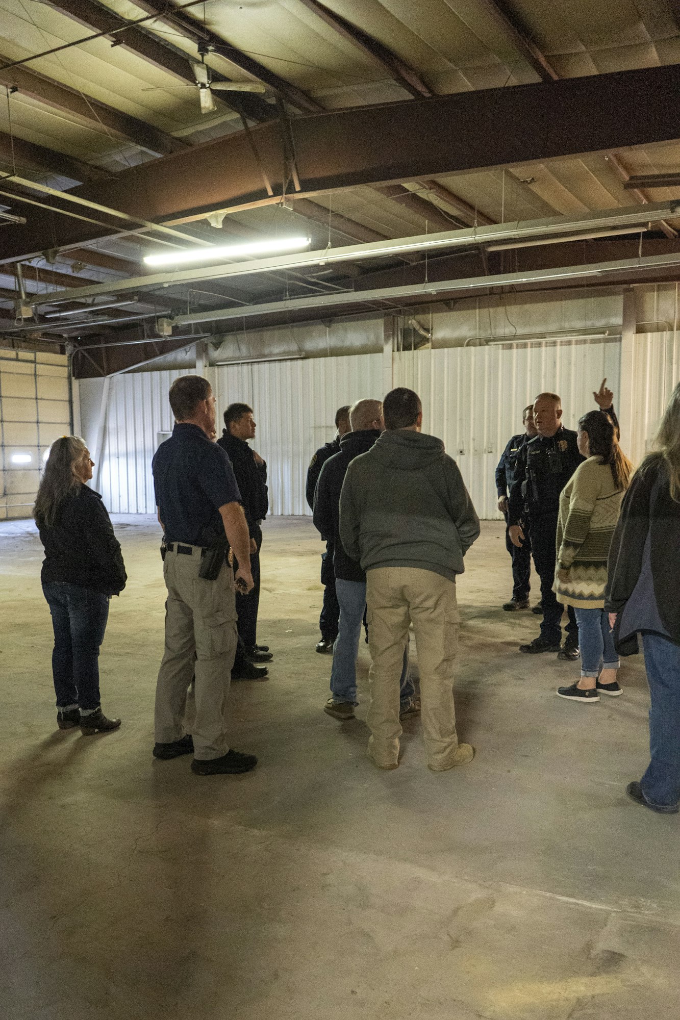A group of people in casual and uniform attire gather in an empty warehouse, with one person gesturing upward.