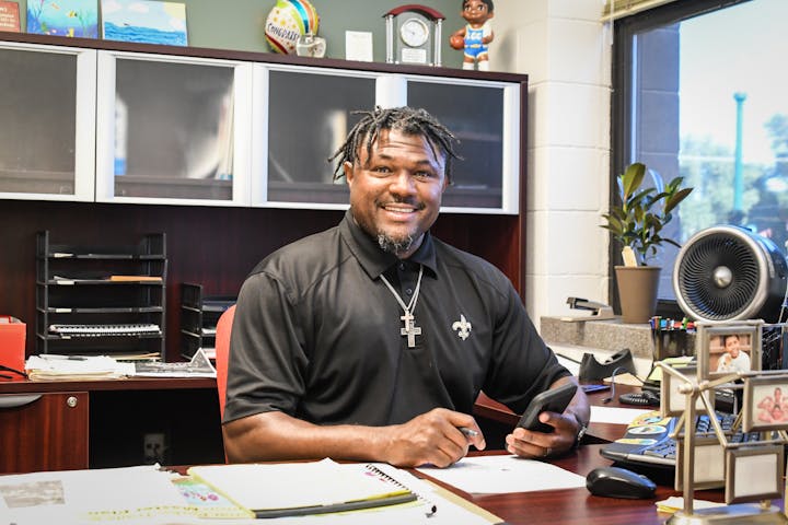 A man in an office, seated at a desk with a phone and papers, smiling at the camera.