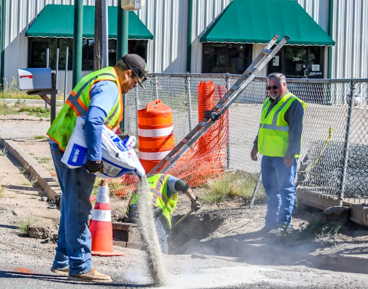 Construction workers in safety vests, one spreading material into a hole, near cones and fencing.
