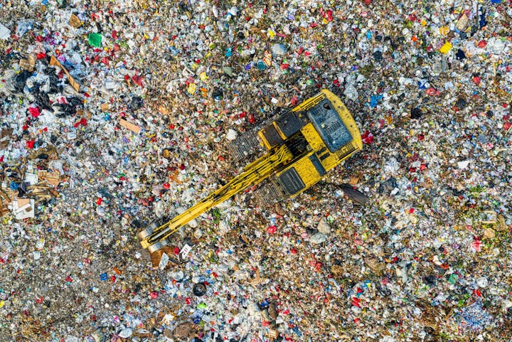 A yellow excavator at a landfill with scattered trash and debris.