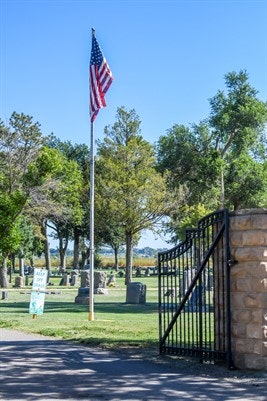 A flagpole with a U.S. flag stands near trees and a cemetery, with an open gate in the foreground.