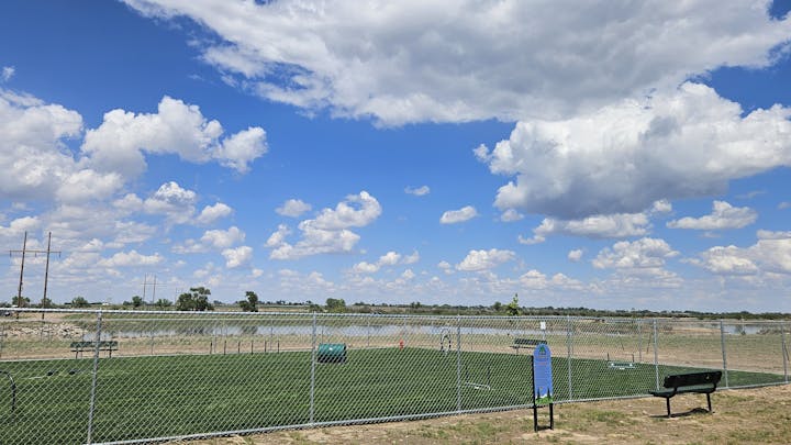 A fenced dog park with exercise equipment, benches, and a clear blue sky with scattered clouds.