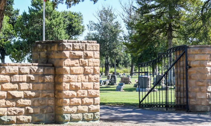 Cemetery entrance with a stone wall and black gate, surrounded by trees and tombstones in the distance.