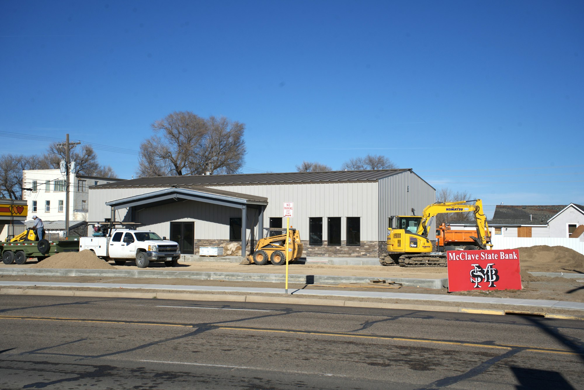 A construction site with a grey building, yellow excavator, white truck, and "McClave State Bank" sign.