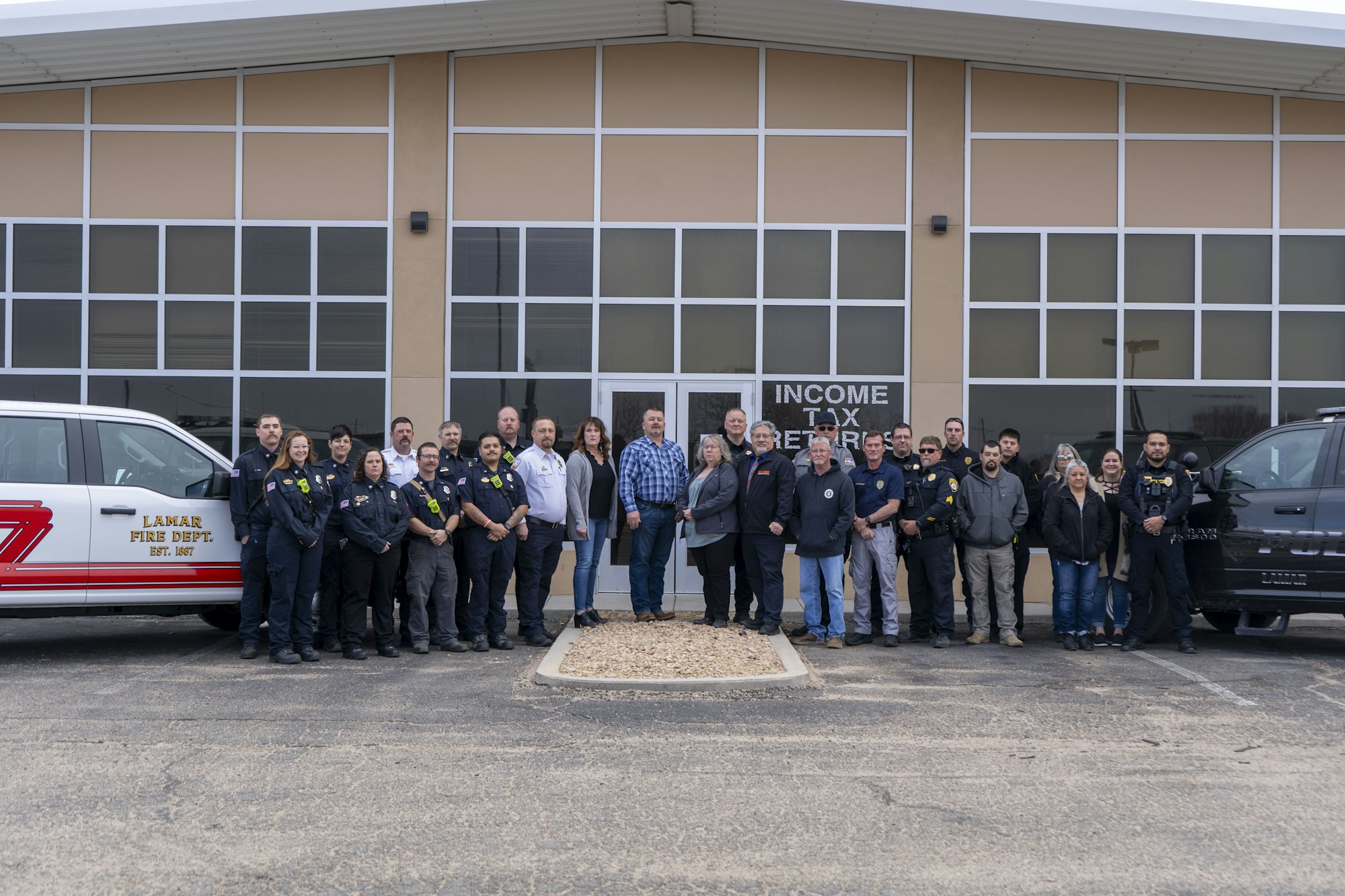 A group of people, including fire department personnel, standing in front of a building labeled "INCOME TAX" with vehicles nearby.