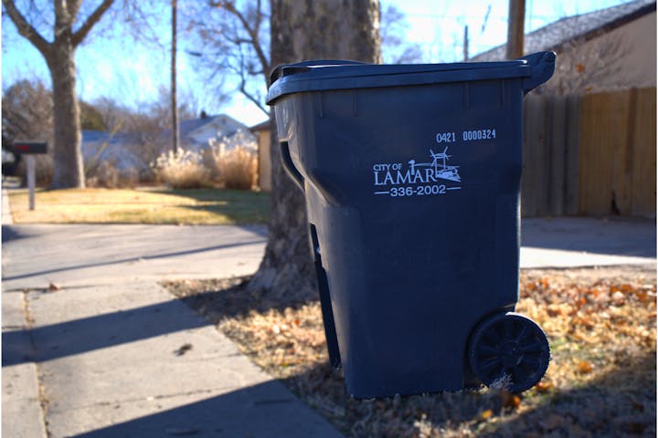 A black trash bin labeled "City of Lamar" on a suburban sidewalk.