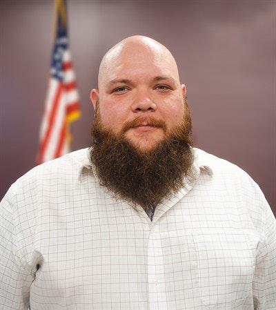 A bearded person in a white shirt with the American flag in the background.