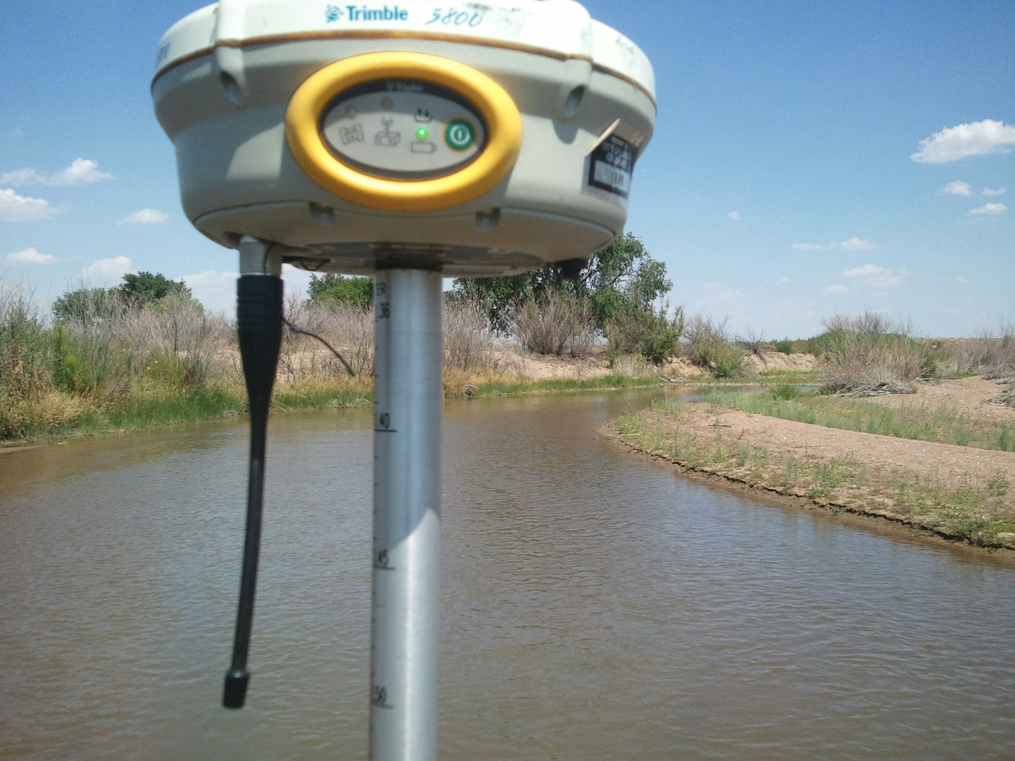 A Trimble GPS device is positioned near a body of water under a clear sky.