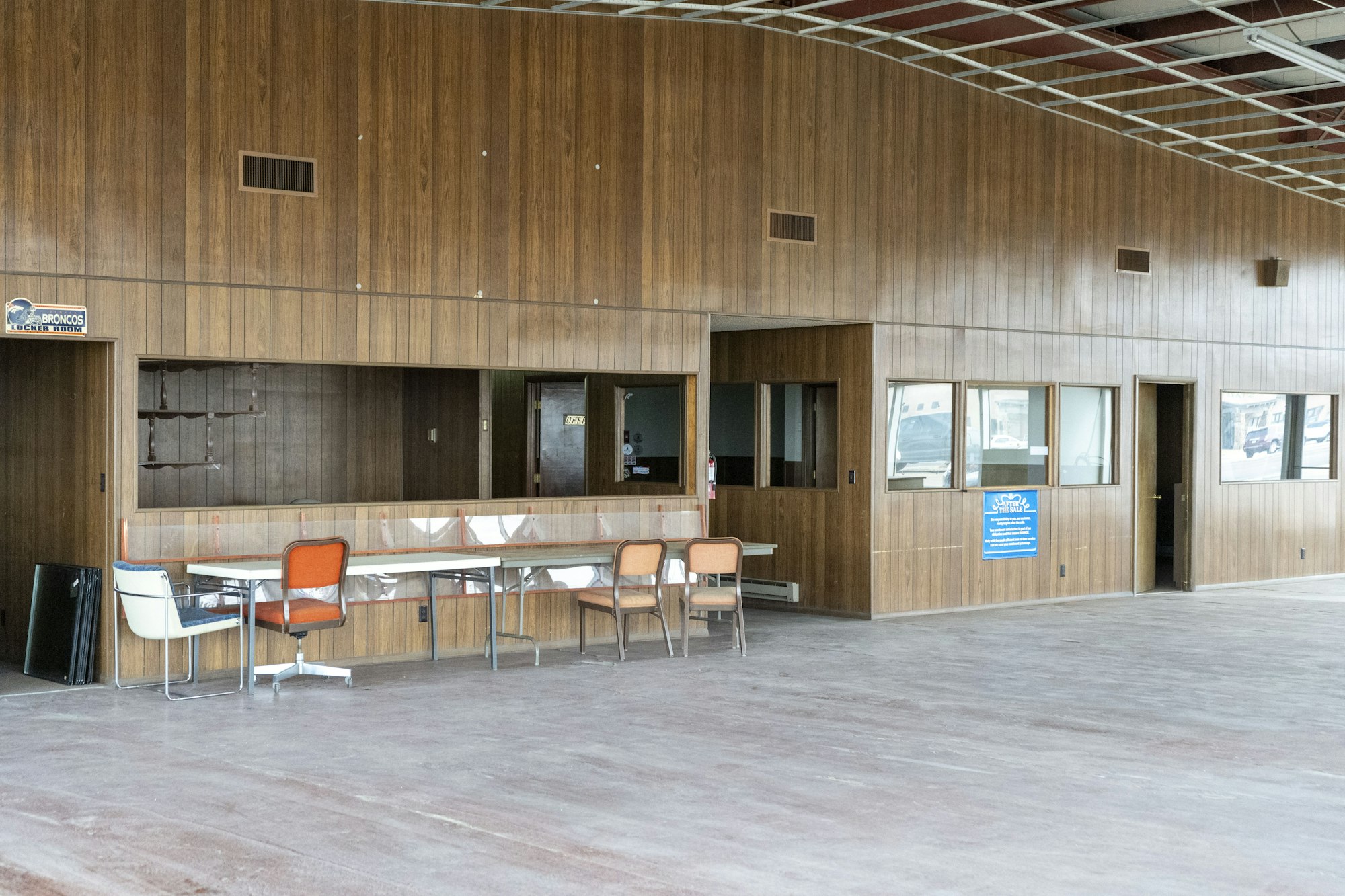 An empty room with wood paneling, chairs, tables, and windows. Signs are visible, and the space appears unused.