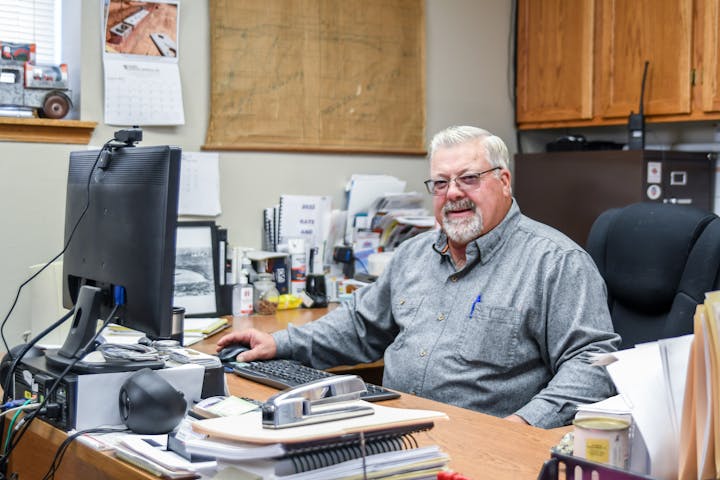 A man sitting in an office at a desk with a computer, surrounded by papers and office supplies.