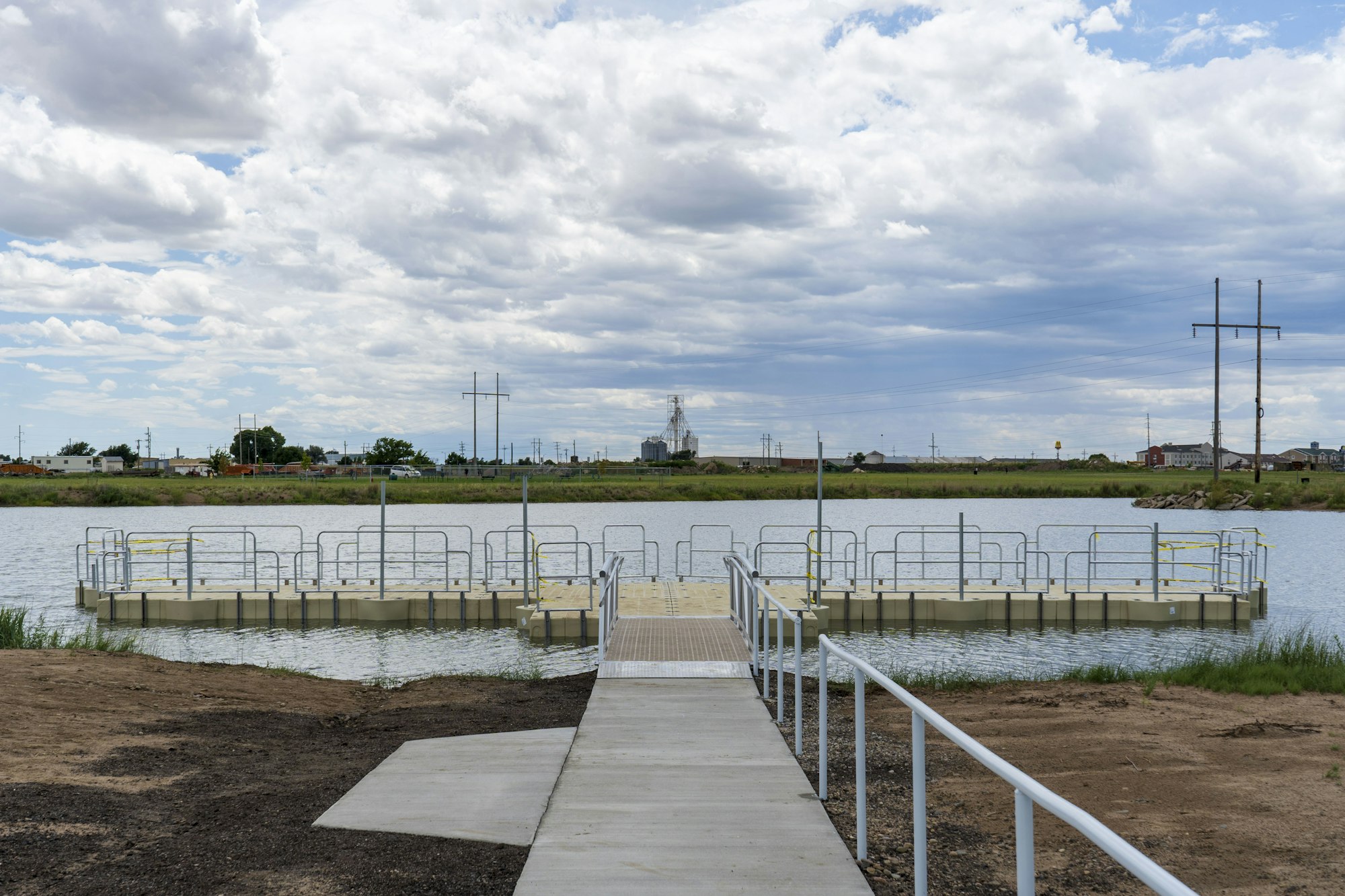 A dock with railings extends over a calm body of water, under a cloudy sky, with buildings and power lines in the background.
