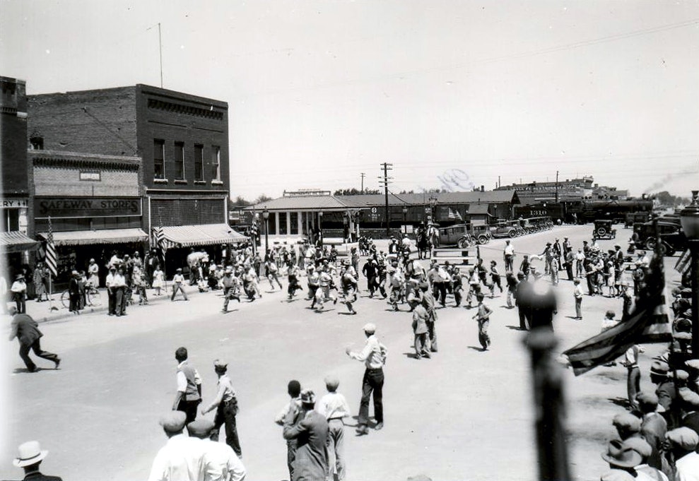 Vintage black-and-white photo of a bustling street scene with people gathered, old cars, and a train in the background.
