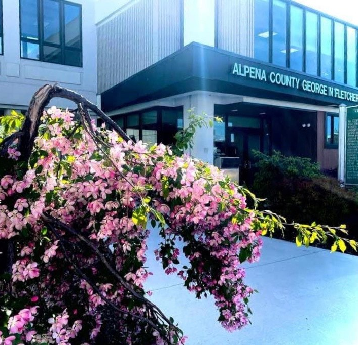 A building named "Alpena County George N. Fletcher" is shown with vibrant pink flowers in front, indicating springtime.
