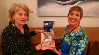Two women are smiling and holding books, including "LitStart" and a GED guide, in a casual setting with a modern background.