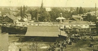 The image shows a historic dock scene with a large boat, buildings, and a crowd of people, likely from the past.