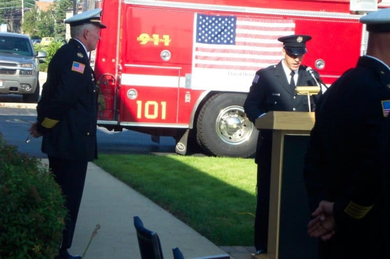 A firefighter in uniform speaks at a podium, with a firetruck displaying an American flag in the background.