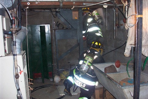 Firefighters in protective gear working in a cluttered basement, using flashlights and equipment.