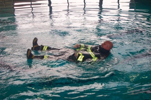 A person in firefighter gear floating on their back in a pool, demonstrating water survival skills.